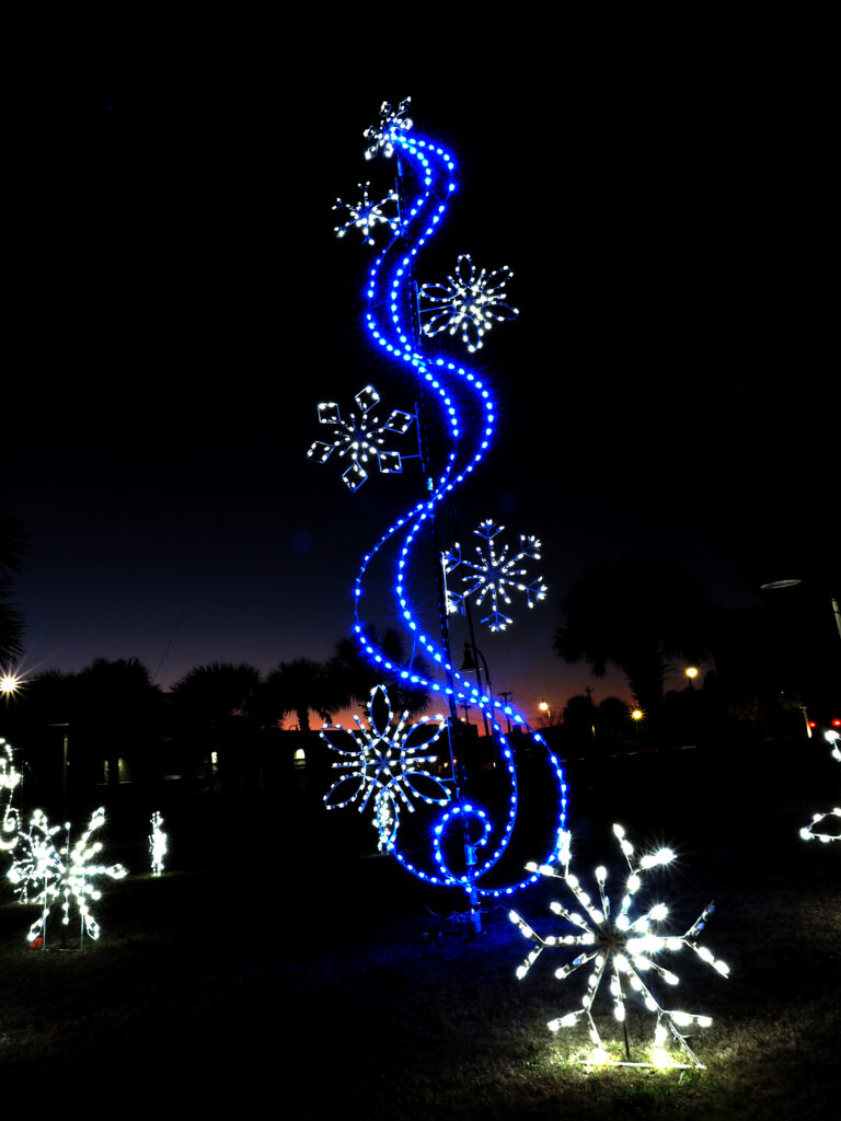 A tall blue snowflake sculpture lit against an orange sunset behind silhouetted palm trees.