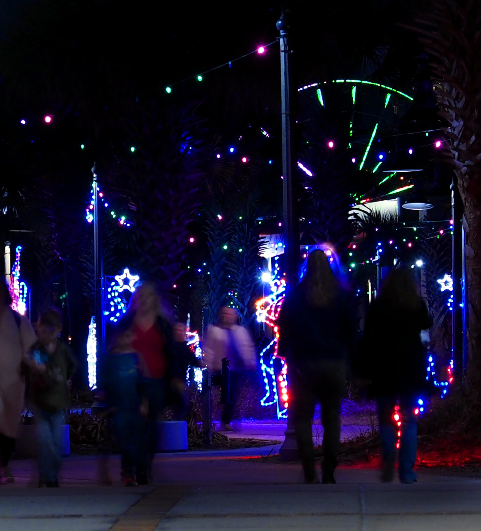 Families walking a palm-lined path strung with colored lights on the Myrtle Beach Boardwalk, the SkyWheel glowing behind them.